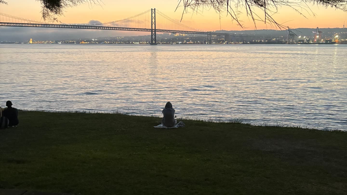 Two people sitting apart on the grass above the Tejo at dusk, the Ponte 25 de Abril across the water