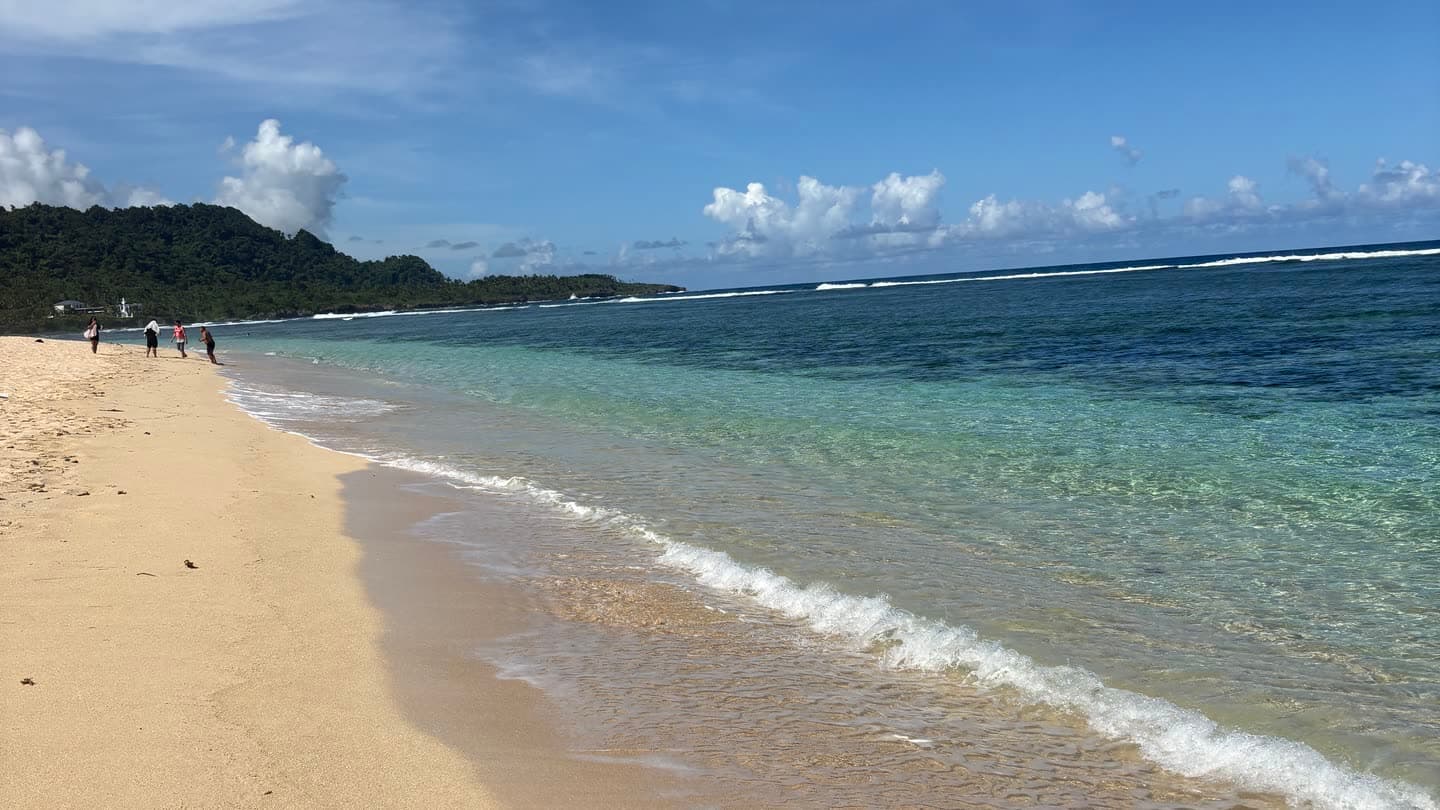 An empty tropical beach with clear shallow water and a reef breaking offshore under a pale sky