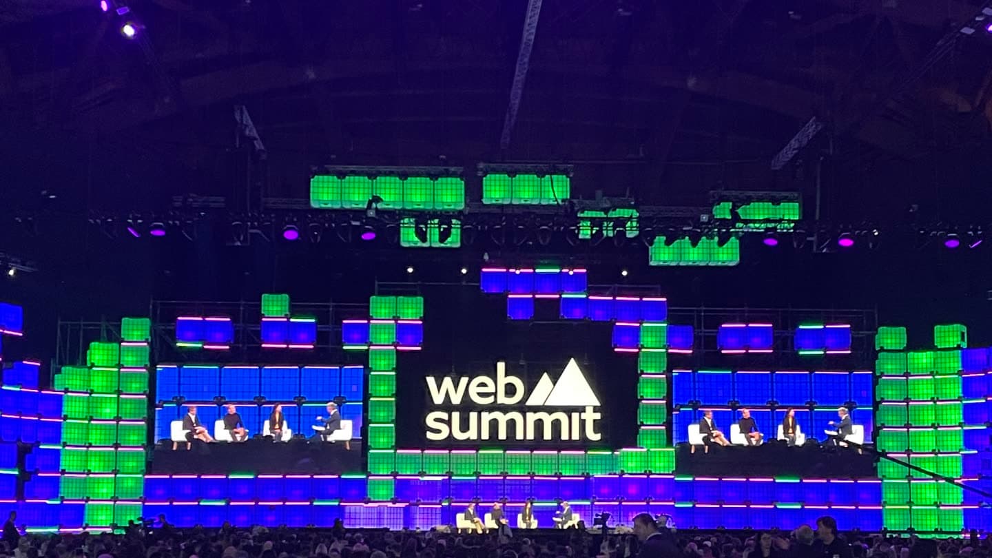 A Web Summit stage in Lisbon seen from the audience, panellists lit against wide blue screens